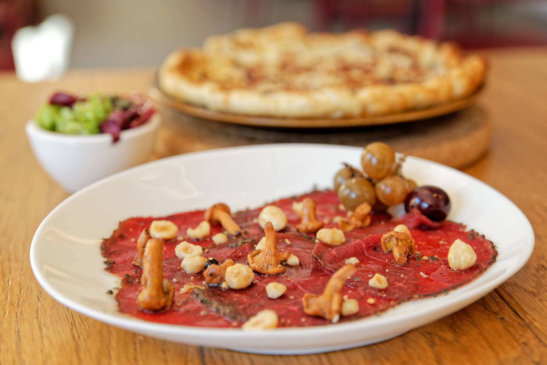 A plate of red meat dish with nuts and olives, a bowl of salad, and a pizza on a wooden table.