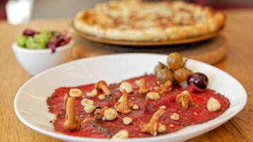 A plate of red meat dish with nuts and olives, a bowl of salad, and a pizza on a wooden table.