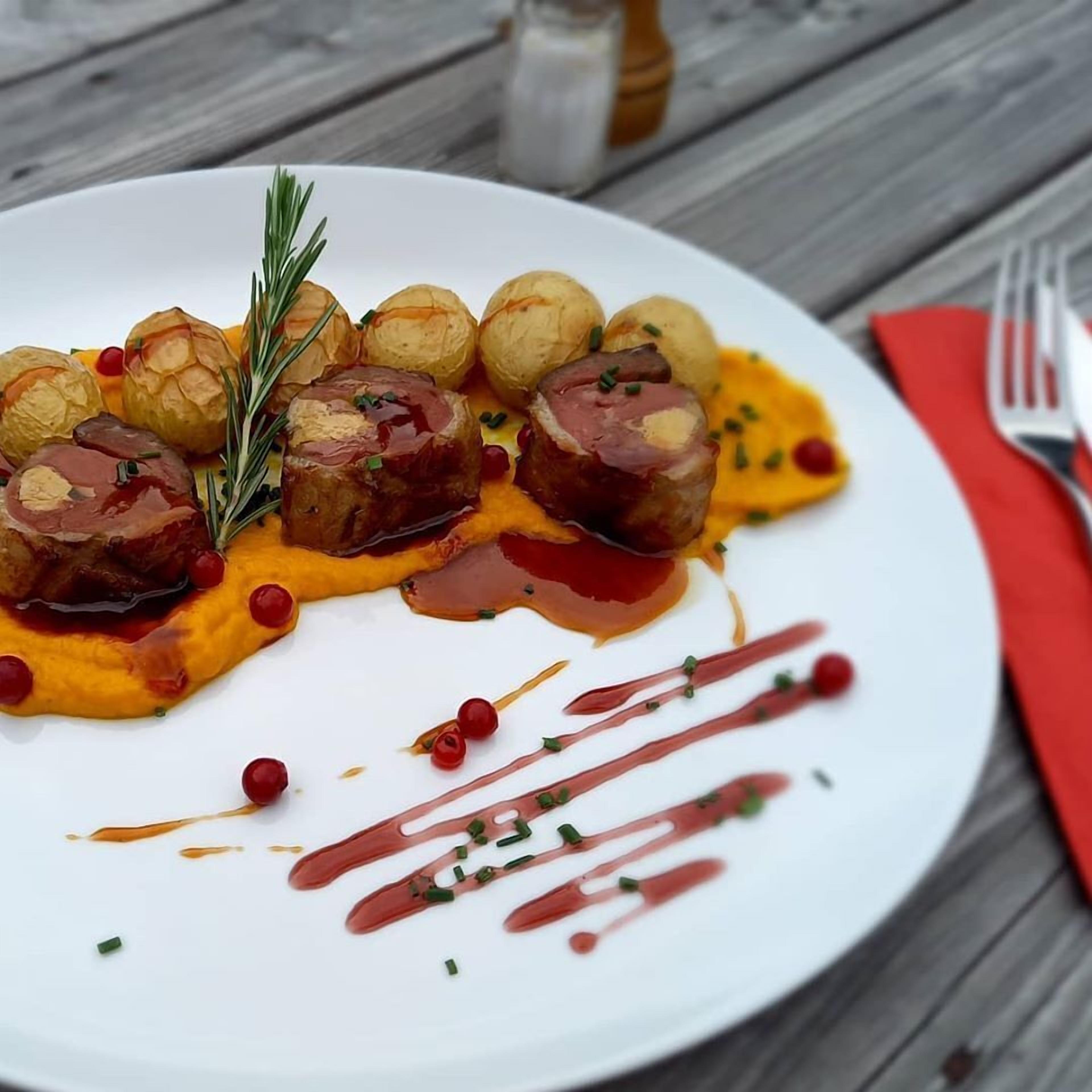 Un plato blanco con patatas asadas, carne y salsa, adornado con hierbas y bayas rojas, sobre una mesa de madera.