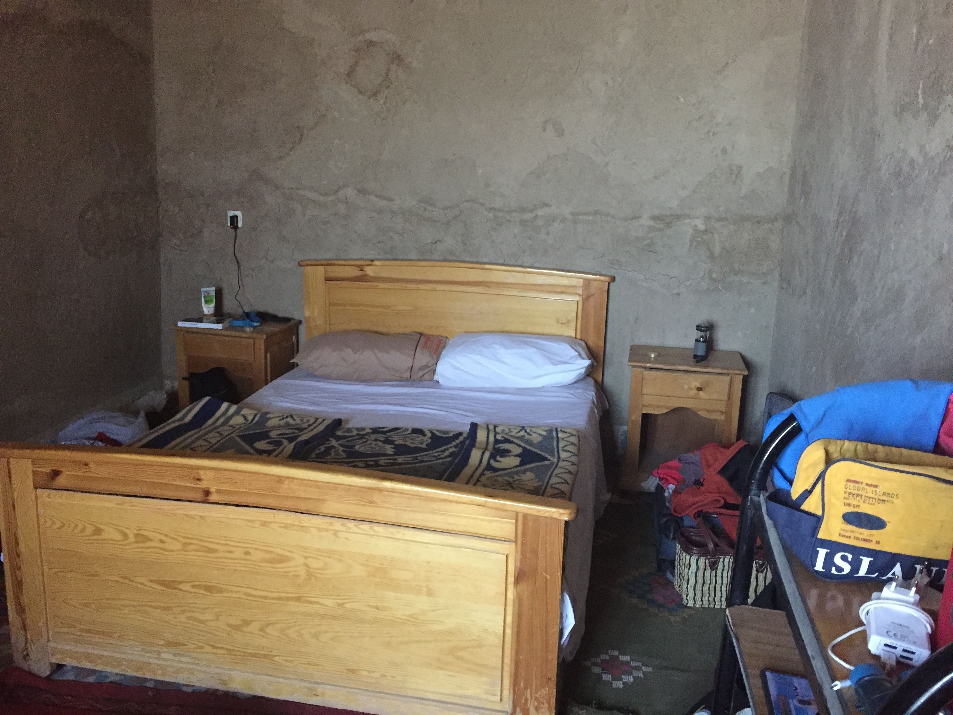 A small, sparsely furnished room with a wooden bed, a nightstand, a small table, and a suitcase.