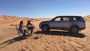 A desert scene with two people at a table with drinks, a car parked nearby, and sand dunes in the background. - Zaid House (Taouz)