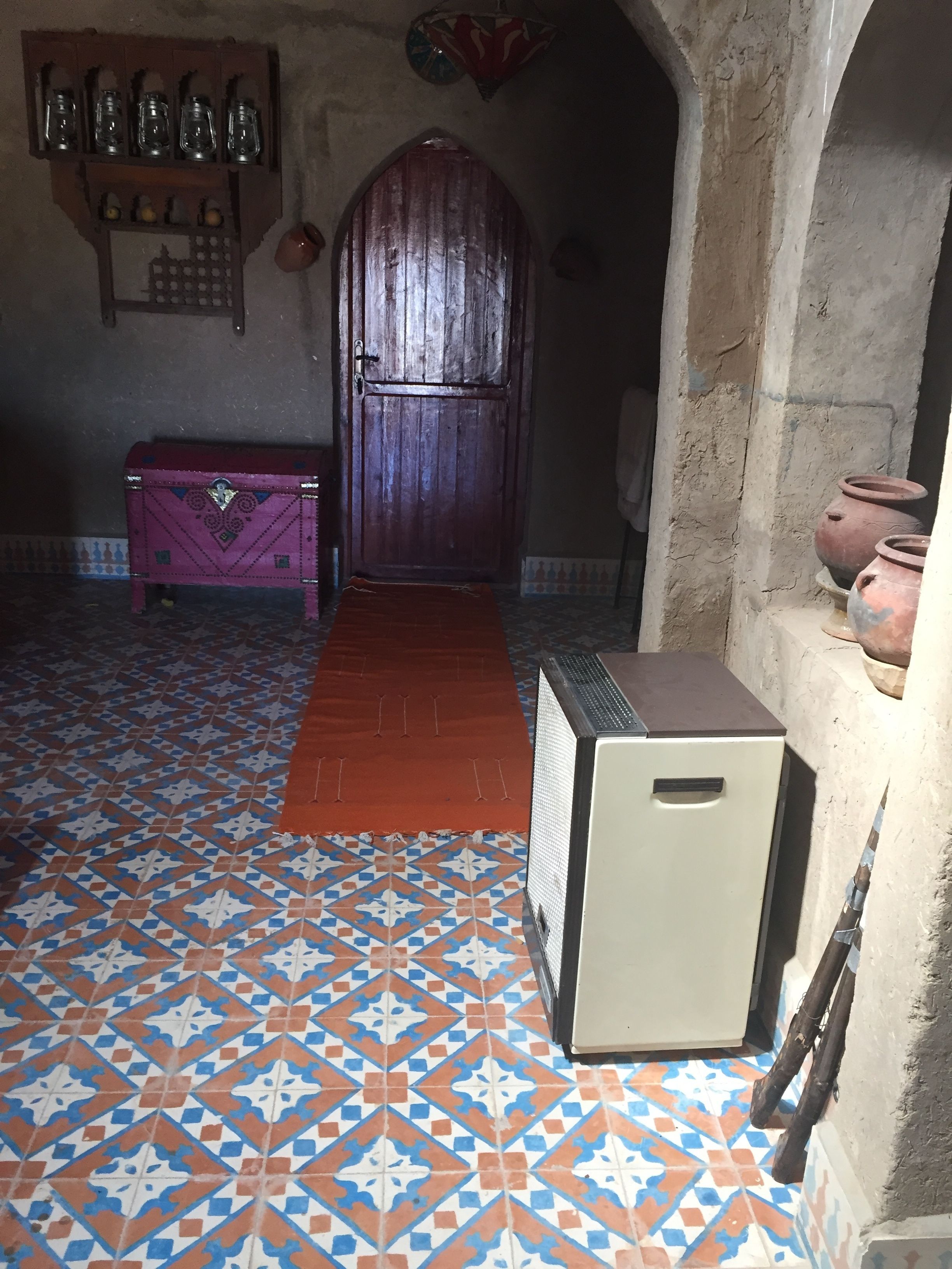 A room with a patterned floor, a wooden door, a small refrigerator, and a shelf with jars.