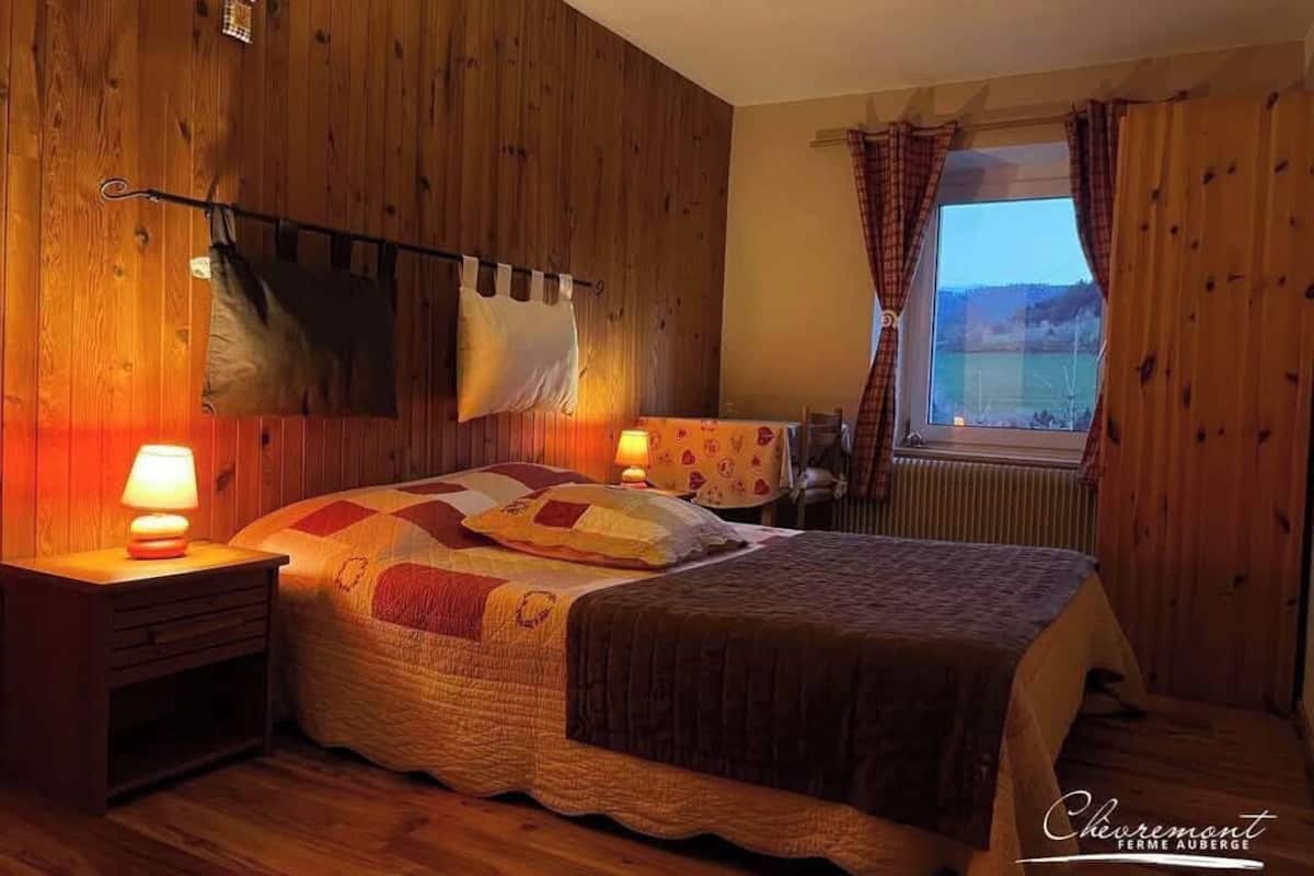 A cozy bedroom with a wooden wall, a bed with a quilt, two bedside lamps, and a window with curtains.