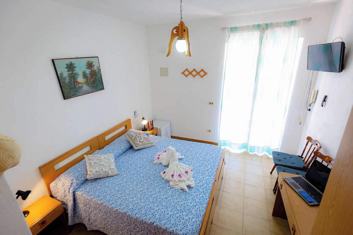 A bedroom with a bed, a desk, a chair, a TV, and a painting on the wall.