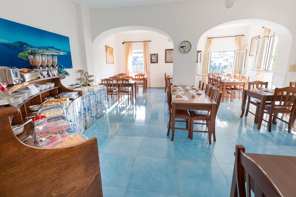 A dining area with multiple tables and chairs, a buffet with various food items, and a large painting on the wall. - Casa Adolfo Ischia (Serrara Fontana)