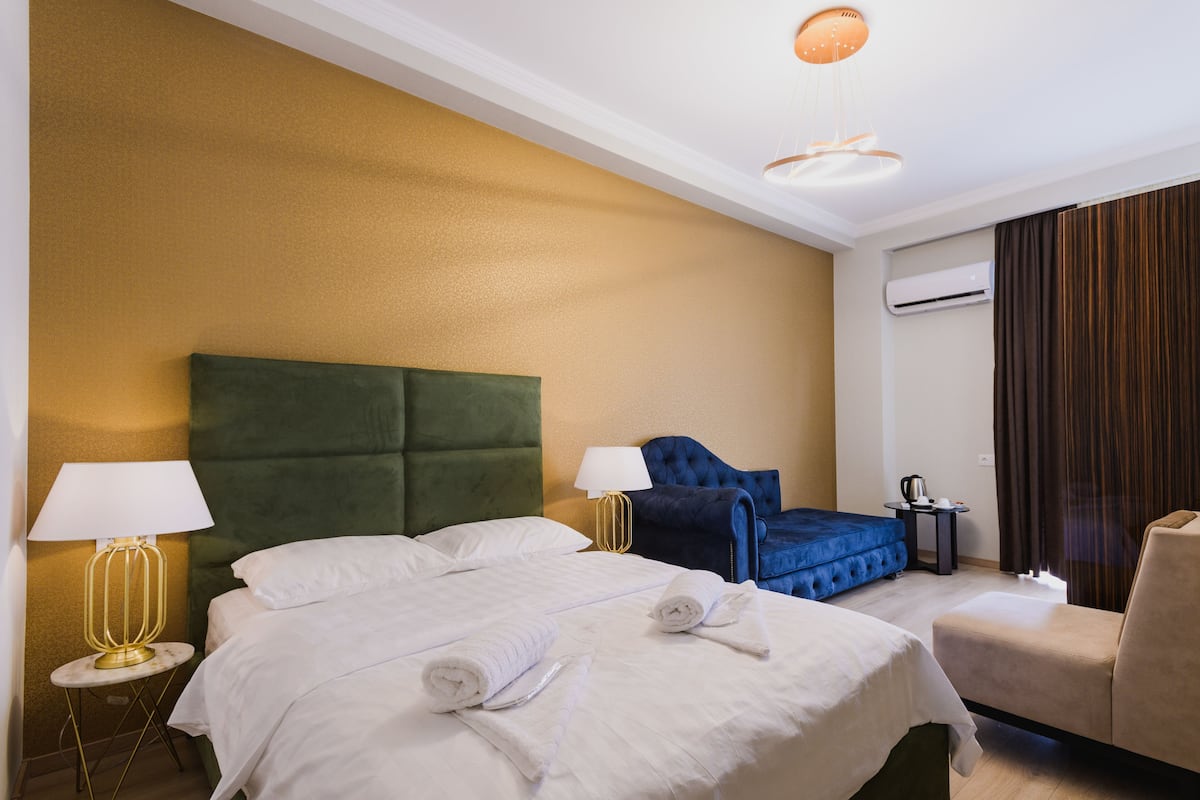 A hotel room with a large bed, a sofa, a desk with a kettle, and a window with curtains.