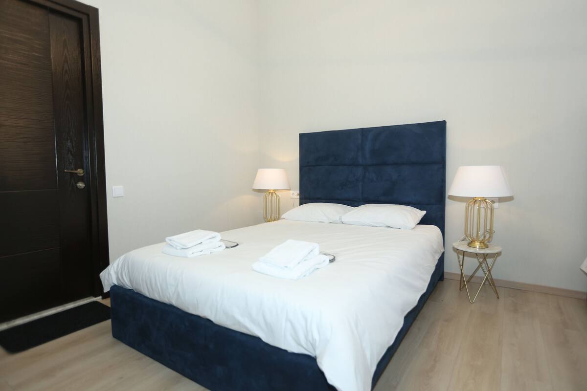 A neatly made bed with white linens, two bedside lamps, and a dark blue headboard in a hotel room.