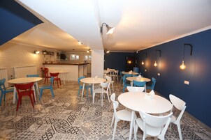 A dining area with round tables and chairs, a kitchen in the background, and patterned flooring.