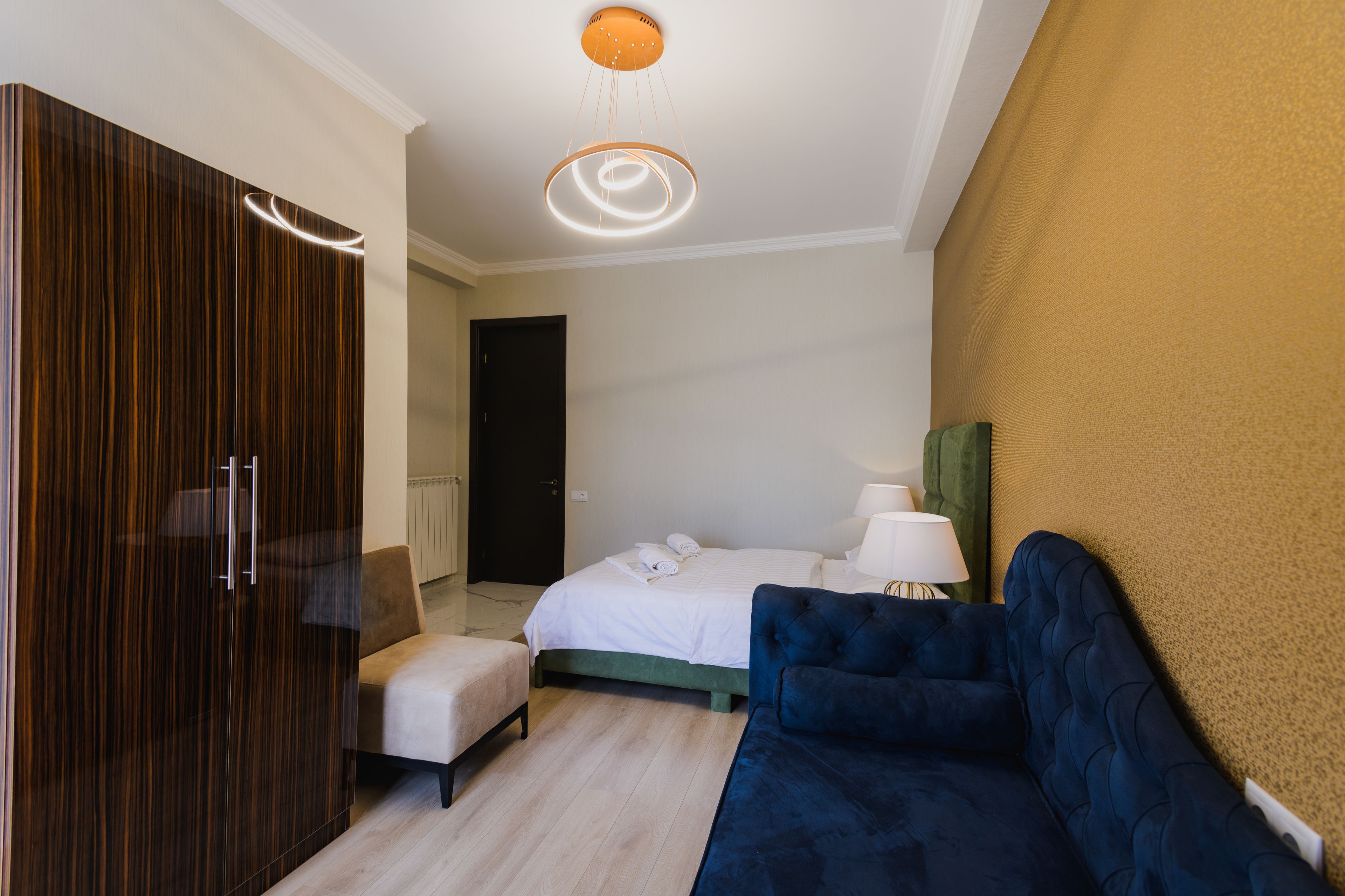 Una habitación de hotel con cama, sofá azul, armario de madera y una mesita con lámpara.