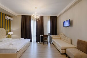 A hotel room with a bed, a sofa, a desk with a chair, a television, and a chandelier.