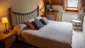 Une chambre à coucher avec une tête de lit en bois, un lit recouvert d’une couverture à motifs, une table de chevet avec une lampe, un bureau en bois, une chaise et une fenêtre donnant sur un paysage.
