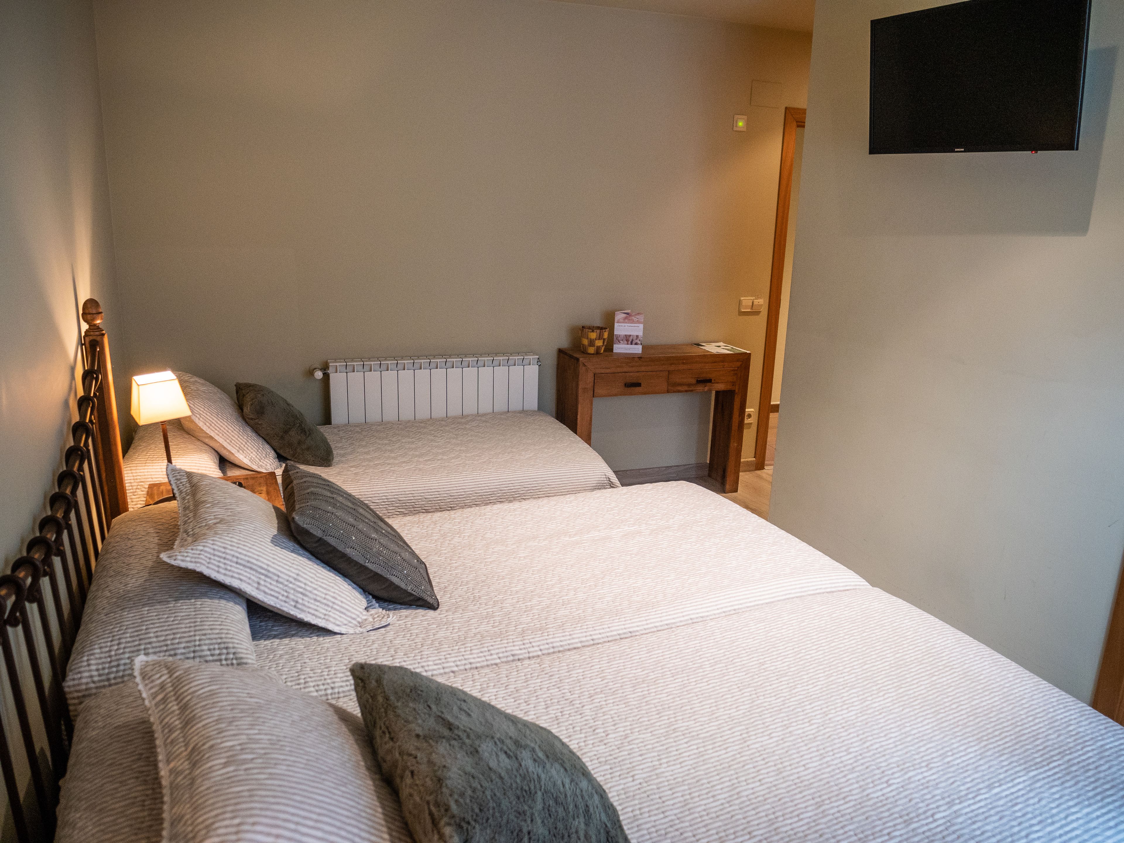 A neatly made bed with a wooden headboard, a bedside table with a lamp, and a small desk with a sign on it.