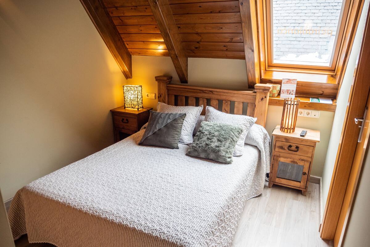 A bedroom with a wooden ceiling, a bed with a white knitted blanket, two pillows, a nightstand with a lamp, and a window with a view of the outside.
