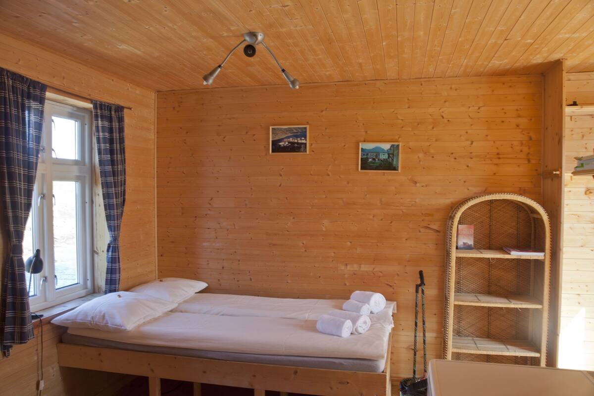 Puisesta materiaalista rakennettu mökkimäinen huone, jossa on sänky, ikkuna verhoilla, hylly ja kattolamppu.