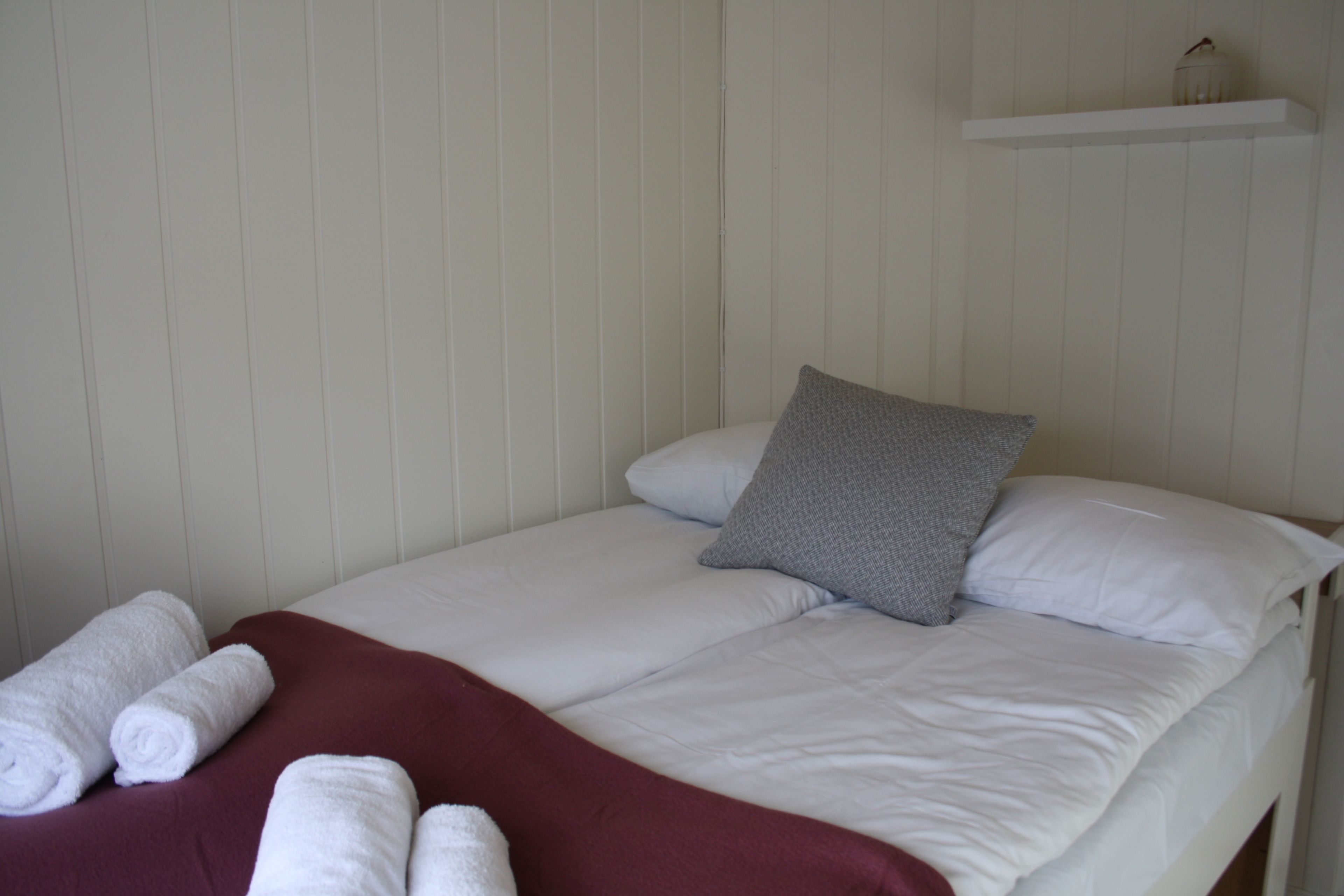 Una cama individual con ropa de cama blanca, un almohadón gris y una manta roja. Hay toallas blancas enrolladas sobre la cama.