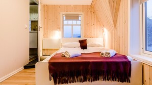 A bedroom with a wooden ceiling, a window, a bed with a purple blanket, and a nightstand with a lamp. - Holmvik Brygge Nyksund (Oksnes)