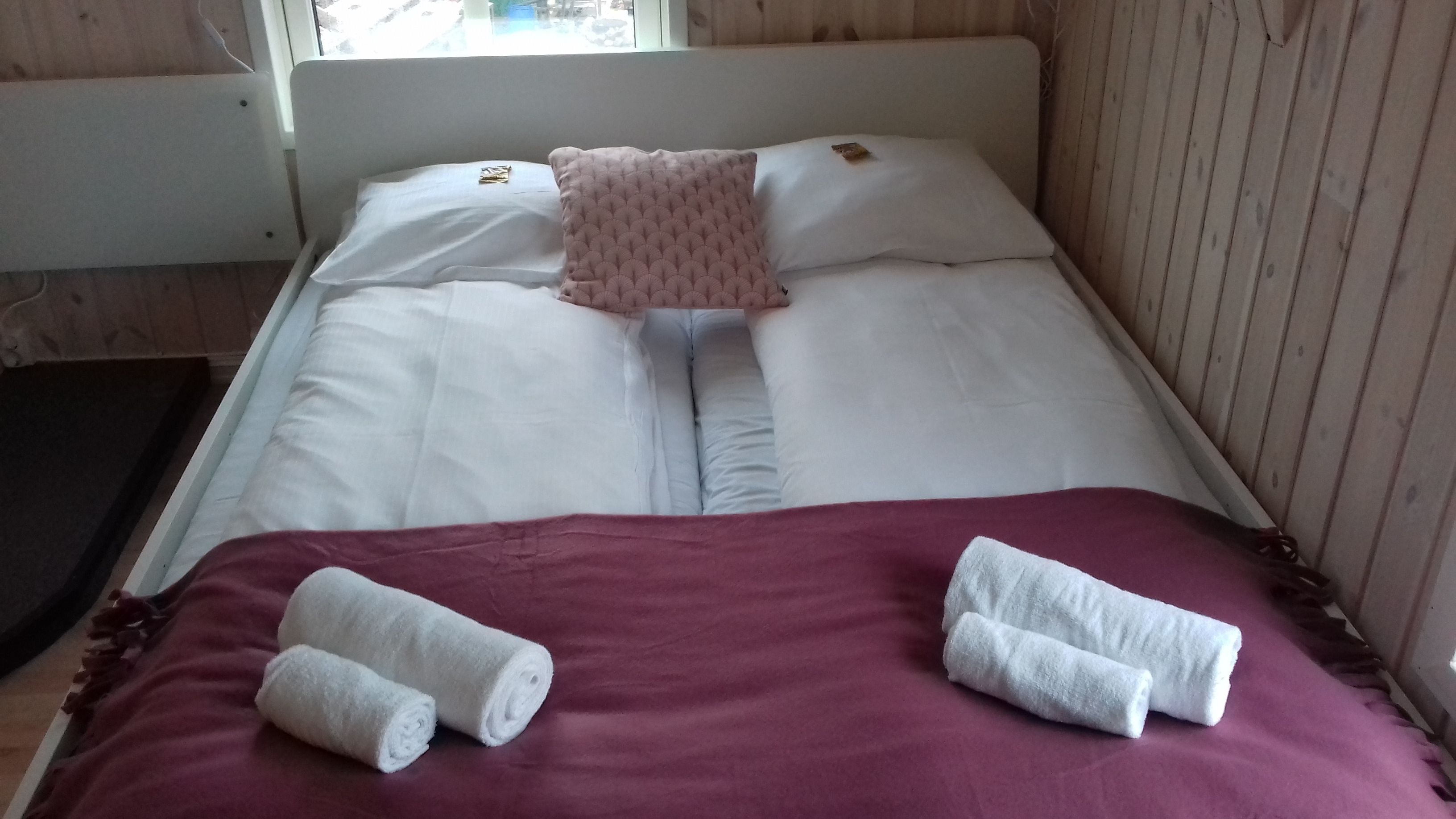 Una cama con sábanas blancas, un almohá de color rosa y dos toallas enrolladas sobre una colcha morada.