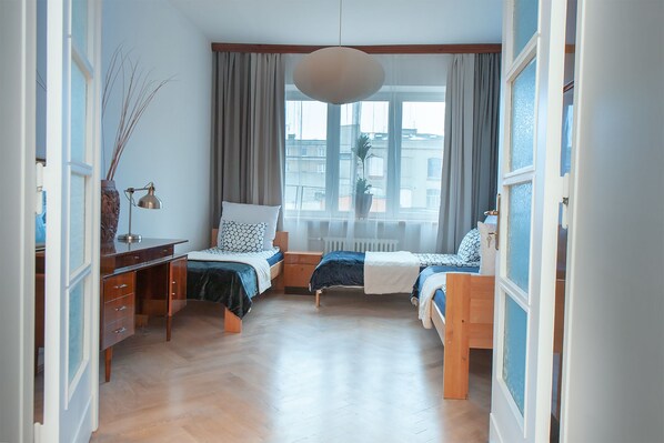 A bedroom with two beds, a desk, a chair, a hanging light fixture, and a window with curtains.