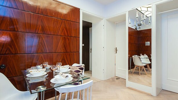 A dining area with a wooden panel wall, a glass dining table, white chairs, and a view into another room with a dining table and chairs. - 4Culture Apart (Lodz)