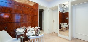 A dining area with a wooden panel wall, a glass dining table, white chairs, and a view into another room with a dining table and chairs.
