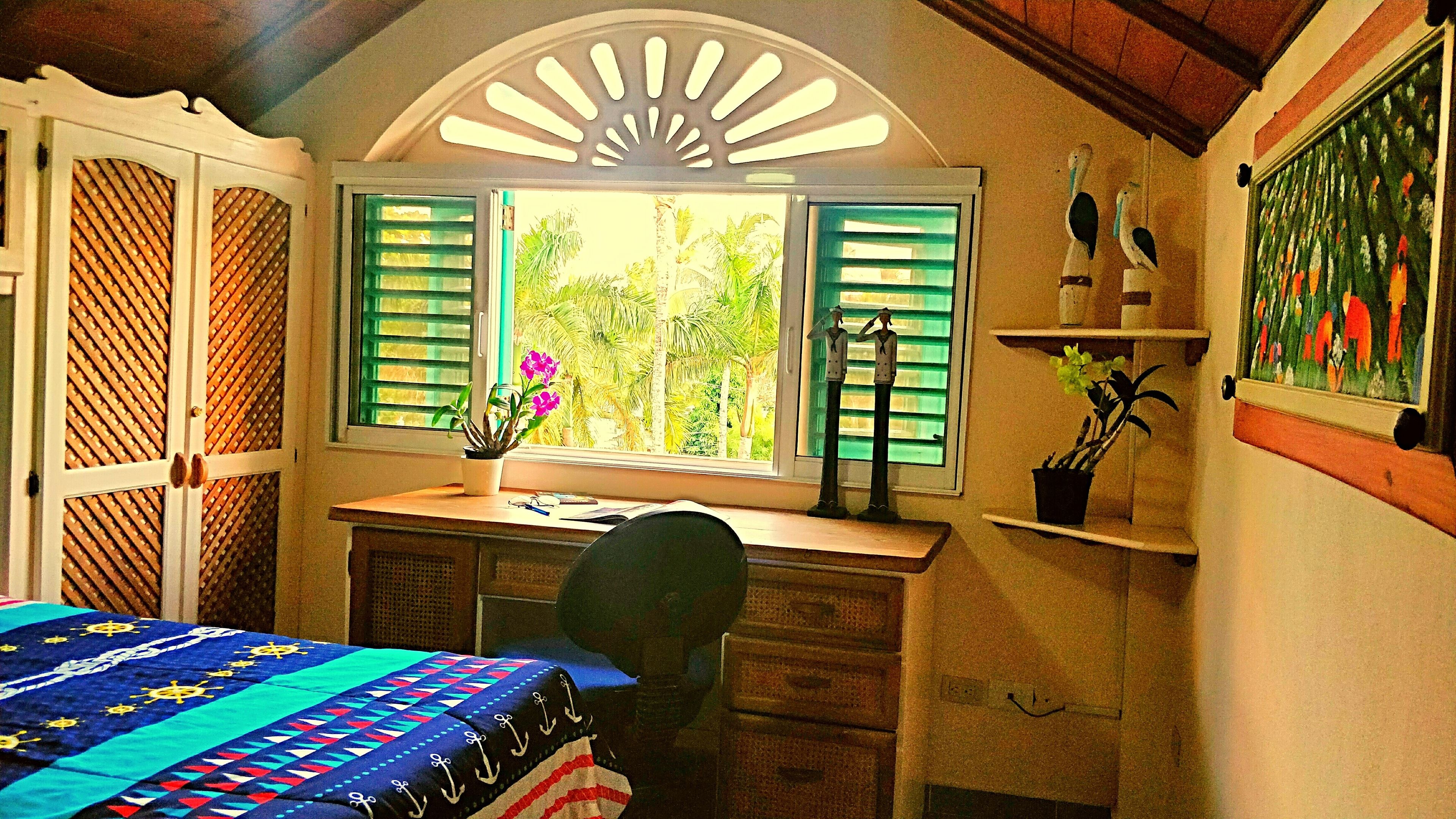 Un dormitorio con cama, un escritorio con computadora, una ventana con contraventanas y un cuadro en la pared.