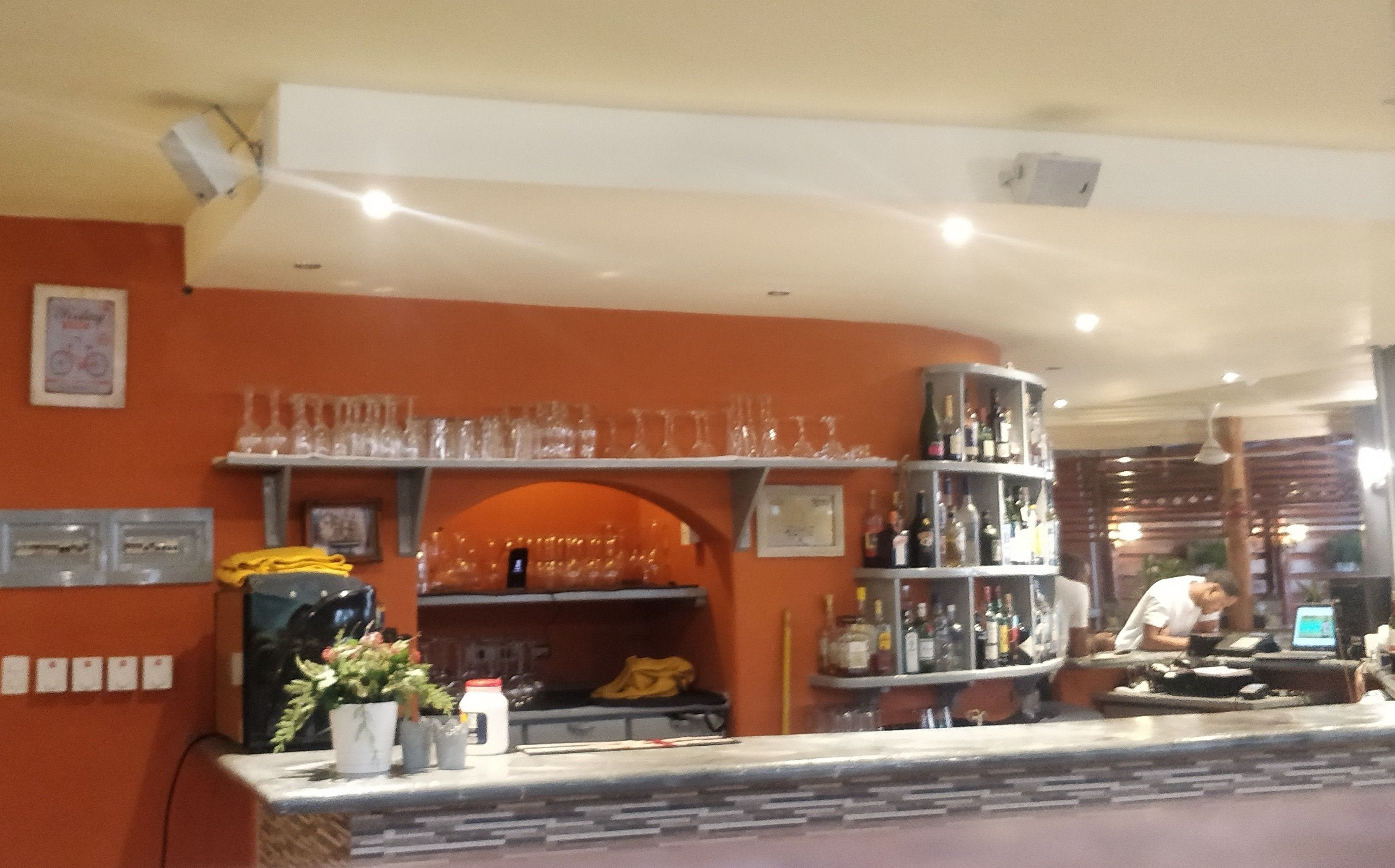 A bar area with shelves of bottles and glasses, a counter with a cash register, and a wall with framed pictures.