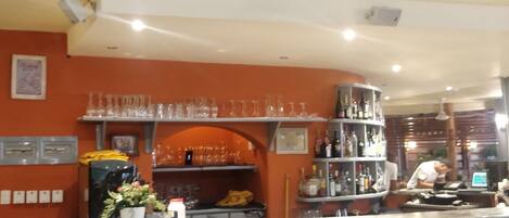 A bar area with shelves of bottles and glasses, a counter with a cash register, and a wall with framed pictures.