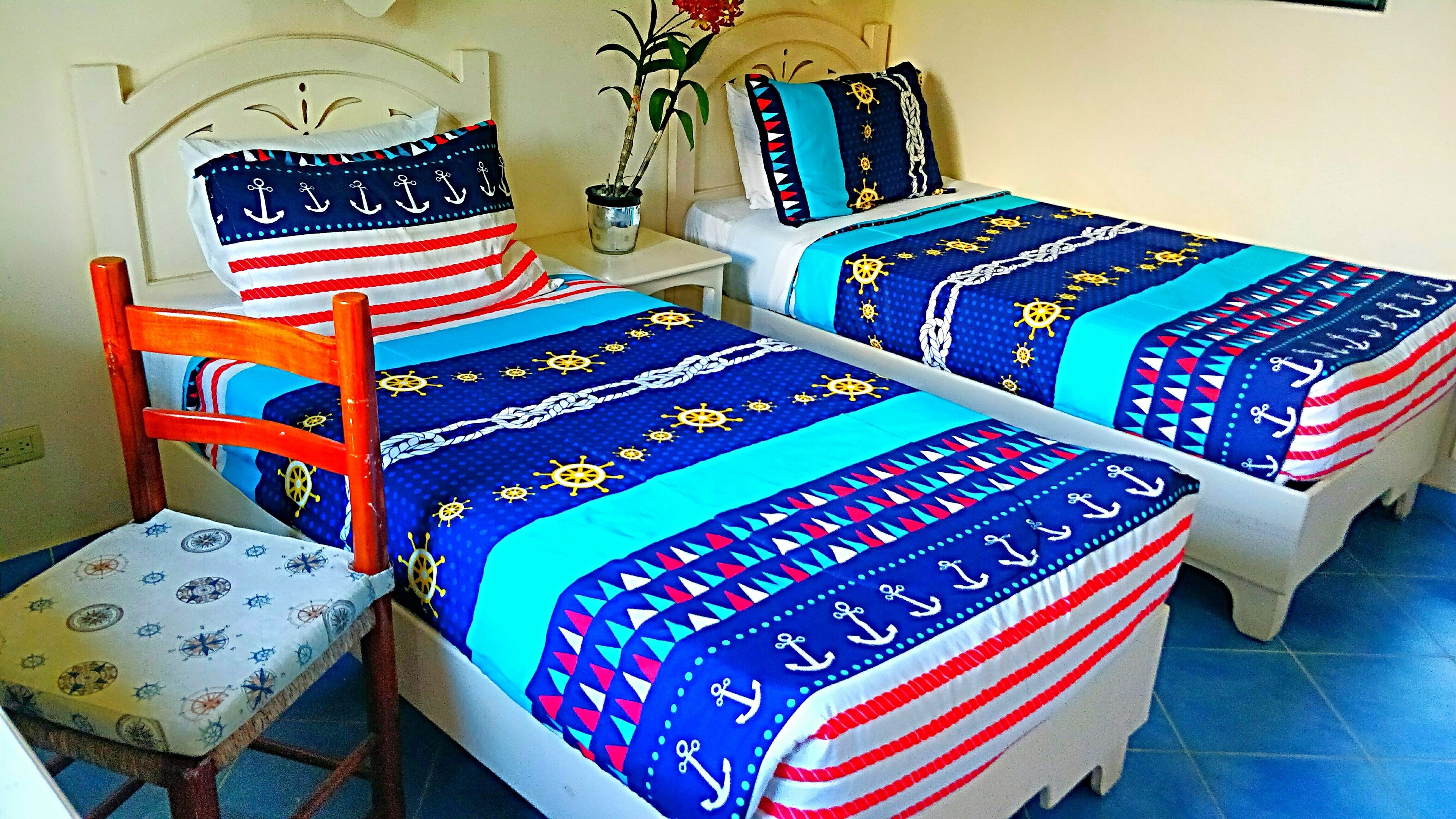 A room with two beds, each with a nautical-themed bedspread, a wooden chair, and a small table with a potted plant.