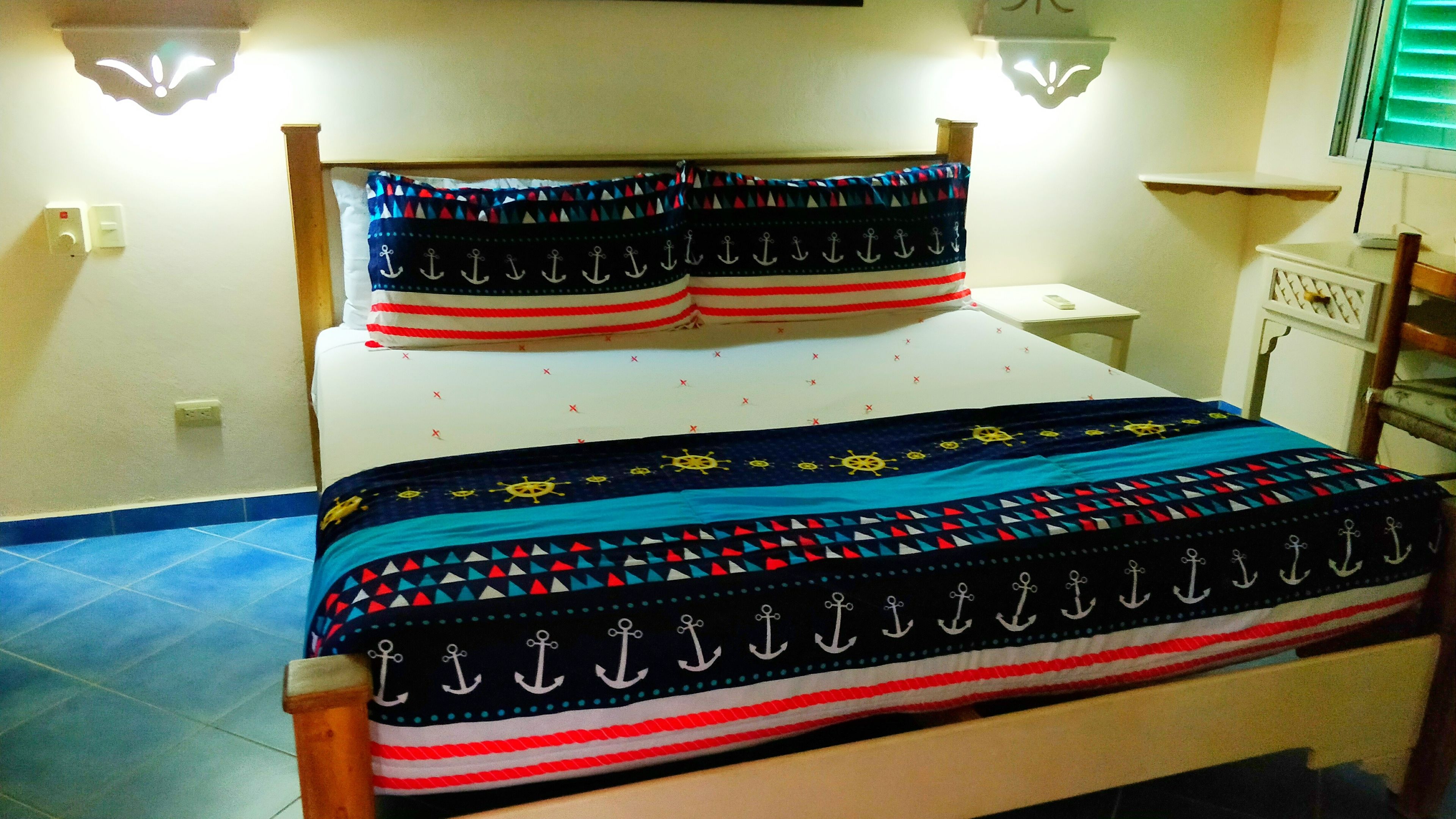 Een bed met een sprei en kussens met een nautisch thema, een nachtkastje met een lamp, en een wandplank met een decoratief object.