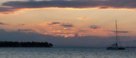 A sunset over a calm sea with a sailboat and a distant shoreline.