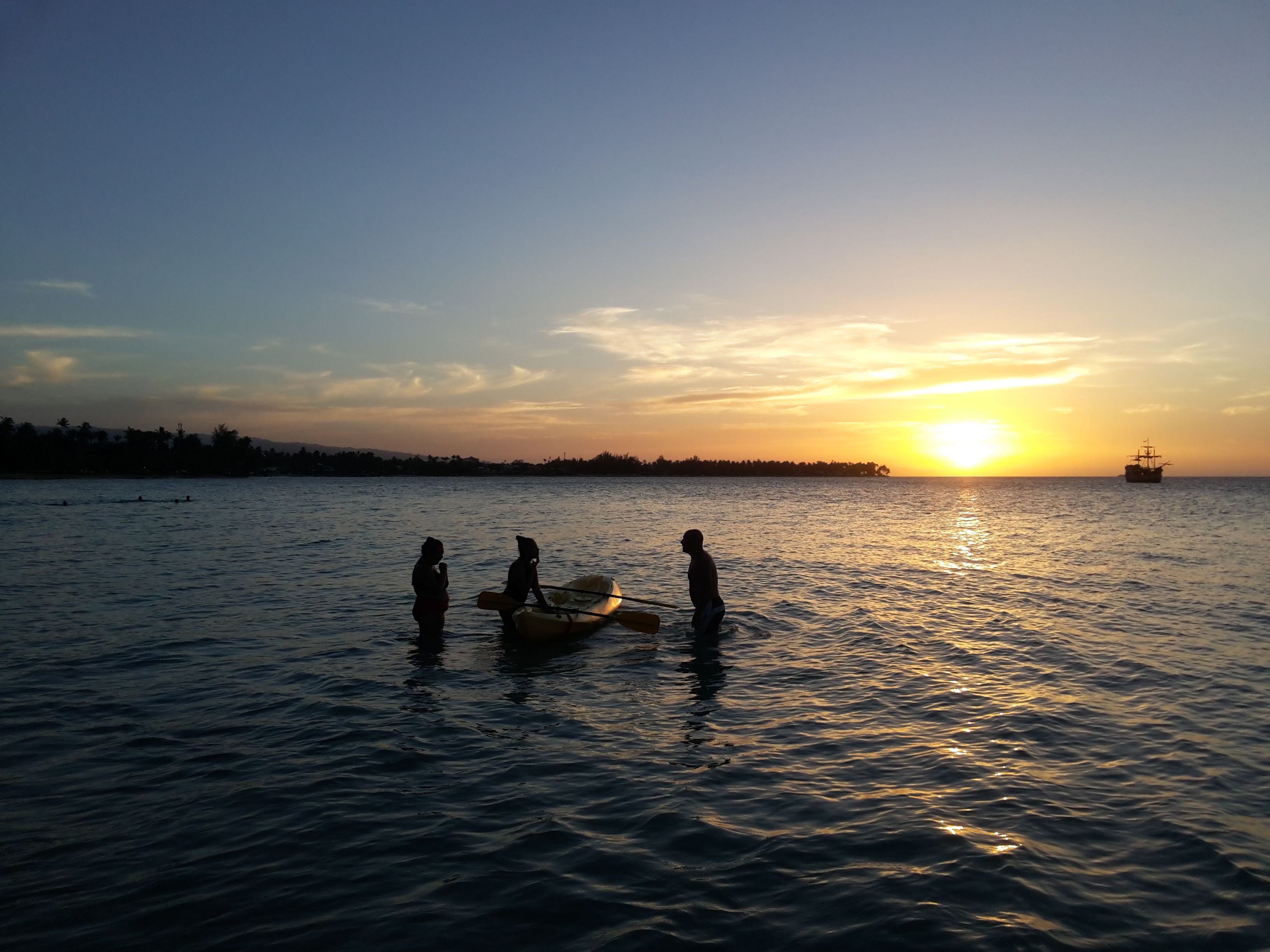 A sunset over a calm sea with three people standing in shallow water, a boat, and a distant ship.