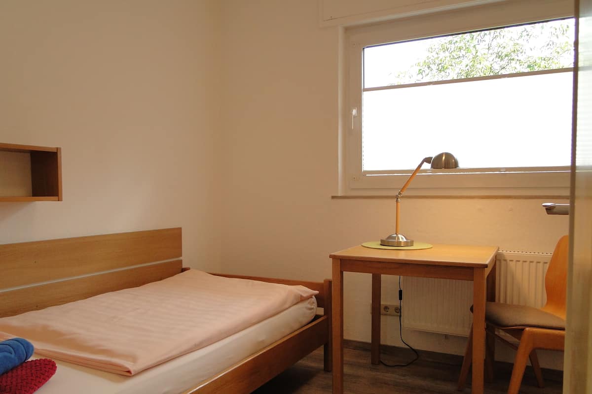 A small, single-bed room with a wooden headboard, a desk with a lamp, a chair, and a window with a view of greenery.