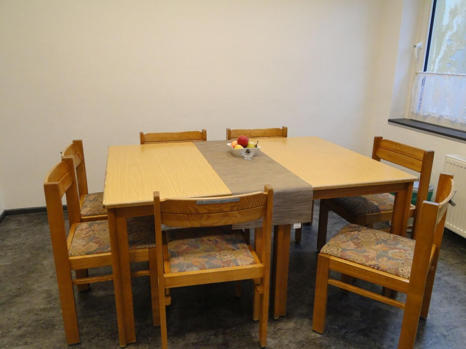 A dining table with six wooden chairs, a central fruit bowl, and a window with blinds.