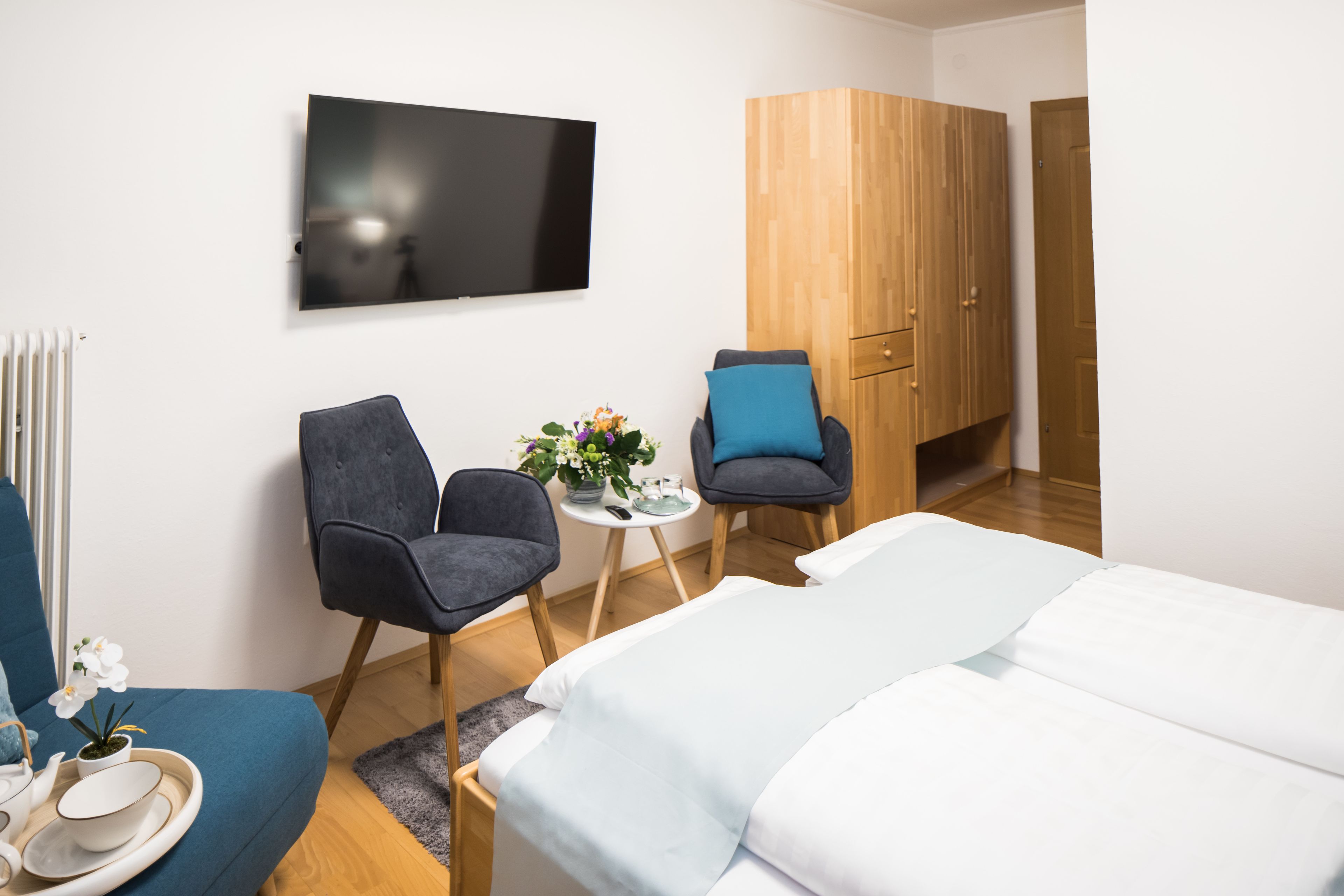Een moderne hotelkamer met een bed, twee fauteuils, een kleine tafel met een bloevas, een flatscreen-tv en een houten garderobe.