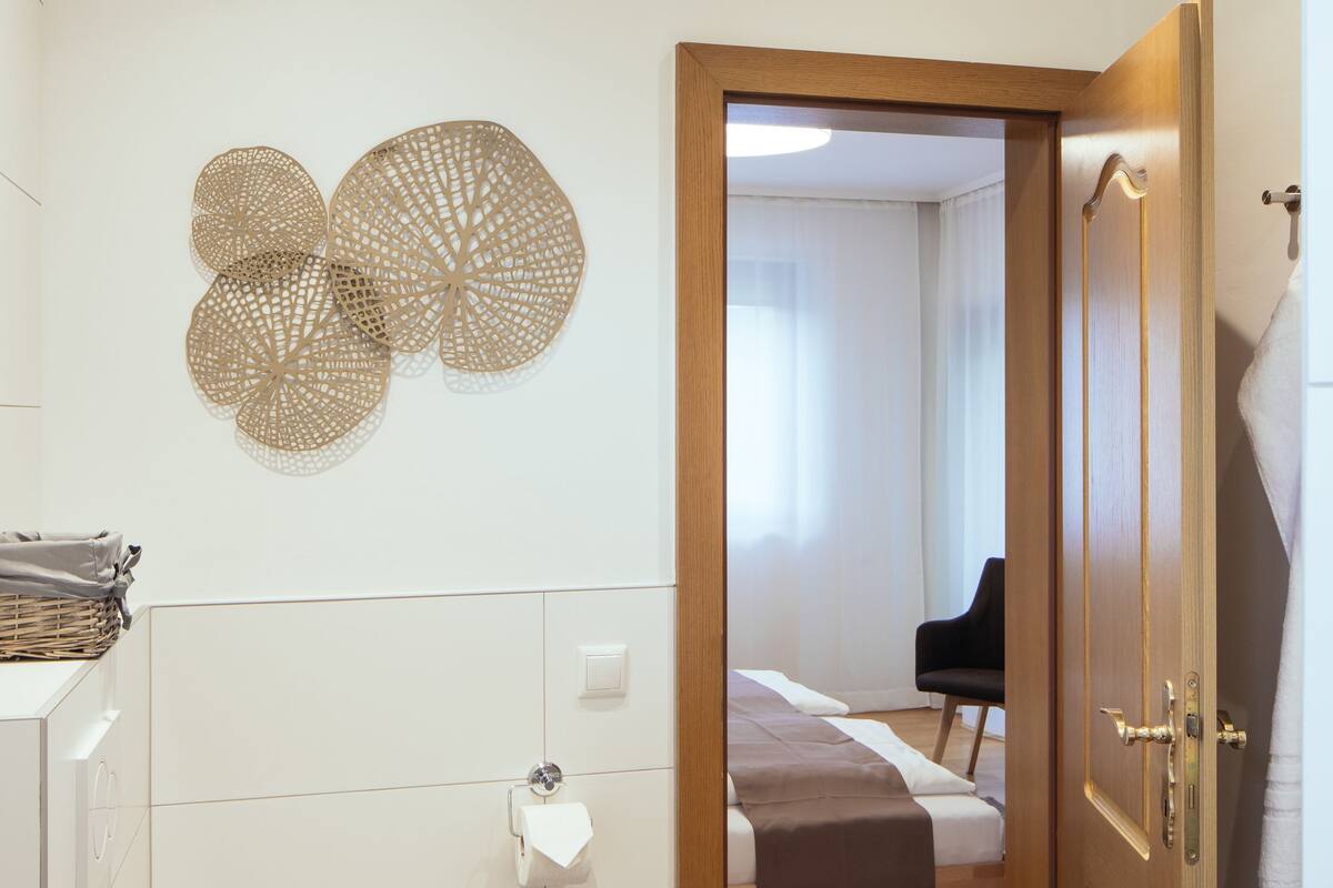 Une salle de bain moderne comprenant des toilettes, un lavabo et un espace douche. Une porte en bois donne accès à une chambre à coucher équipée d’un lit et d’une chaise.