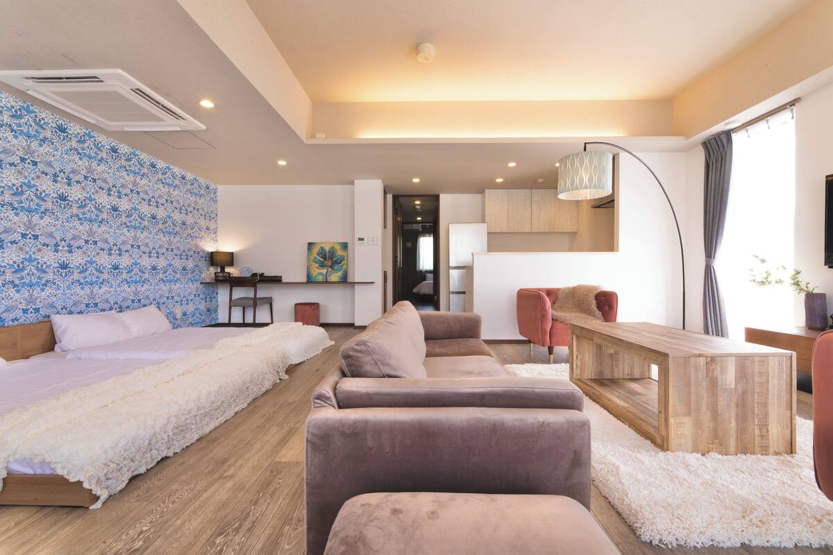A modern hotel room with a large bed, a sofa, a wooden coffee table, and a kitchen area in the background.