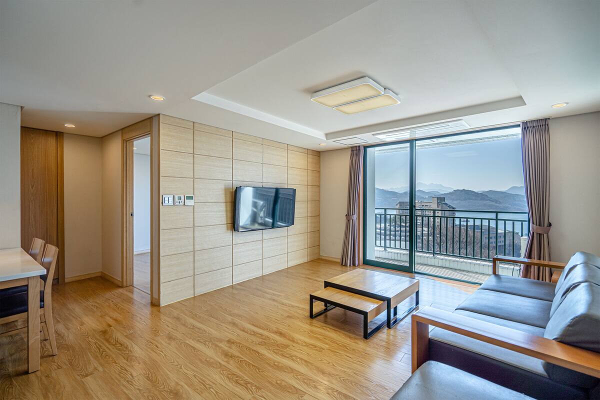 A modern living room with a wooden floor, a flat-screen TV mounted on the wall, a sofa, a coffee table, and a dining area with a table and chairs.