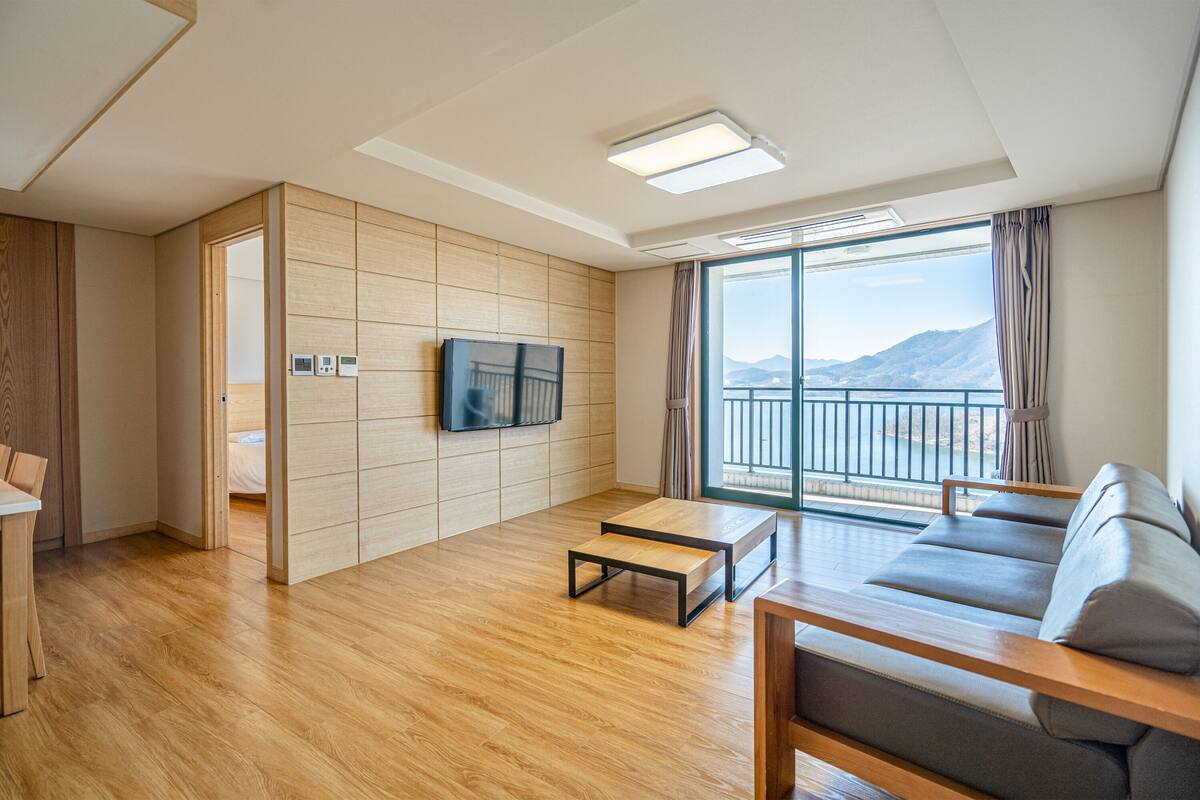 A modern living room with a wooden floor, a sofa, a coffee table, a TV mounted on the wall, and a large window with a view of the mountains.