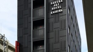 Um edifício moderno com fachada em vermelho e preto marcante, apresentando um grande logotipo de flor e o nome "Sakura Sky Hotel Kashiwa".