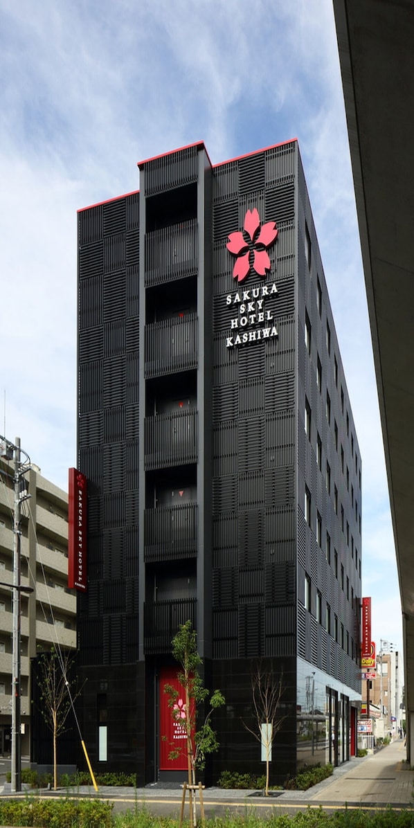 Un edificio moderno con una fachada llamativa de rojo y negro, que presenta un gran logotipo floral y el nombre "Sakura Sky Hotel Kashiwa".