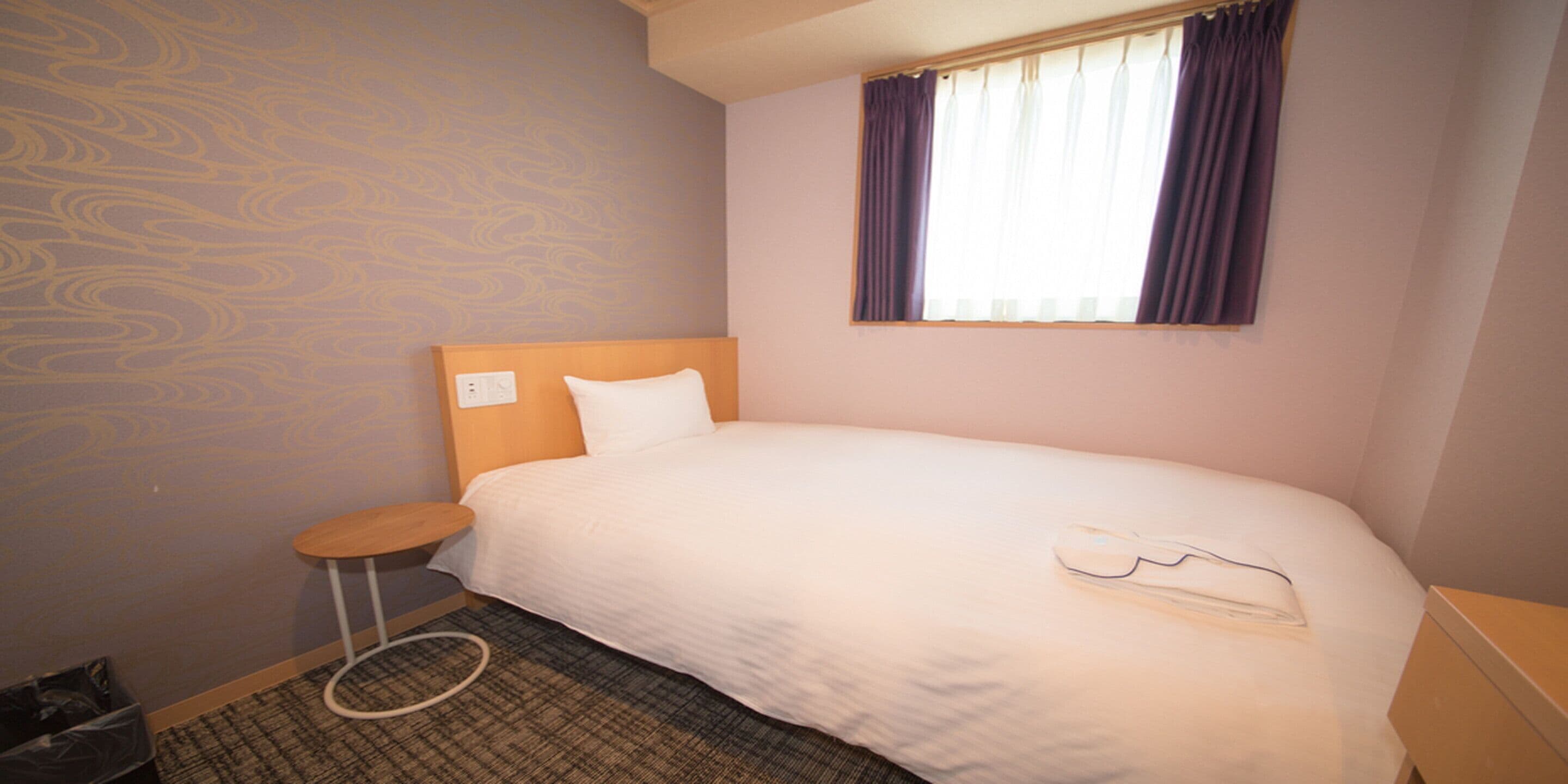 Habitación de hotel con una cama individual, una mesita redonda pequeña, un cesto de basura y una ventana con cortinas.