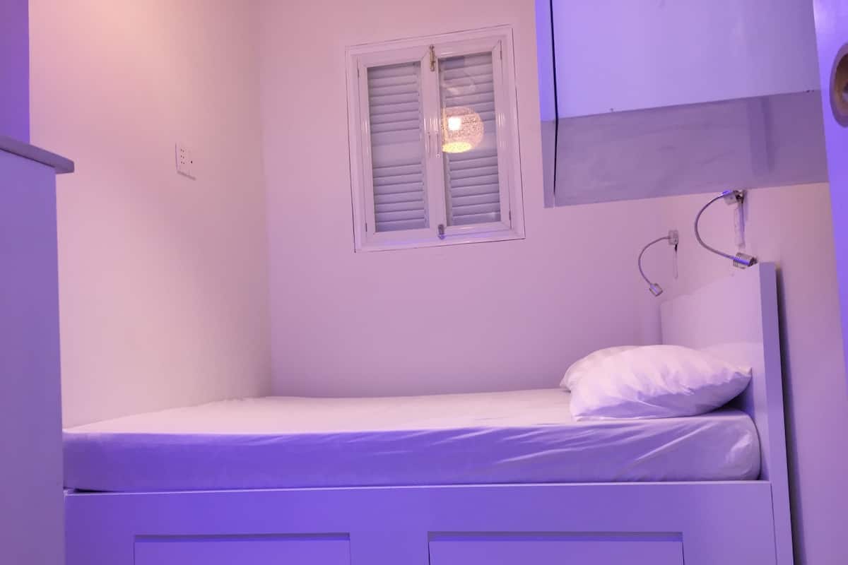 A small, single-bed room with a window, a bedside lamp, and a power outlet on the wall.