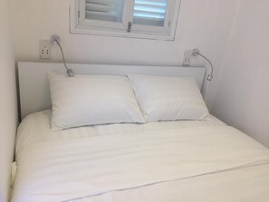 Una cama con ropa de cama blanca y dos almohadas, una ventana con persianas y enchufes en la pared.