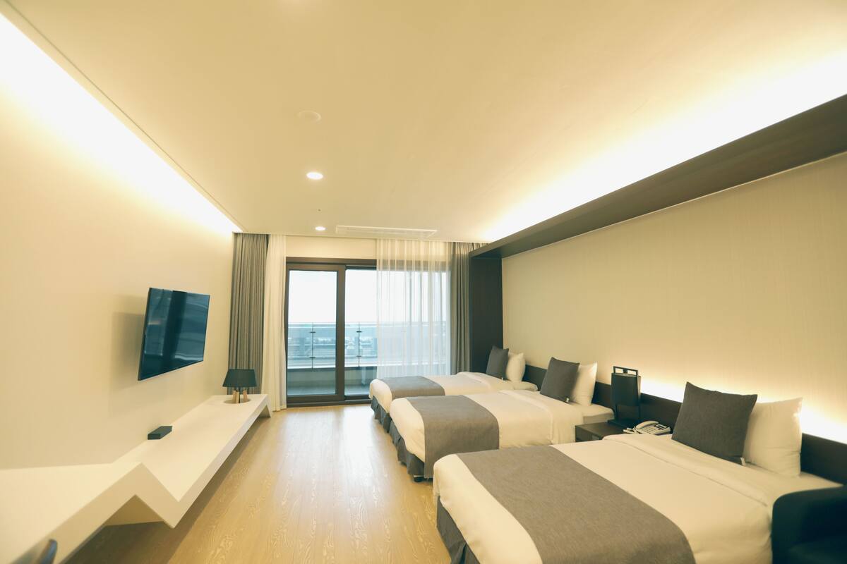 A hotel room with two beds, a flat-screen TV, a desk, and a view of the city through the window.