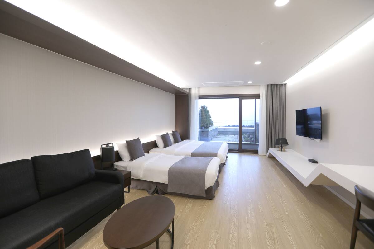 A modern hotel room with a sofa, two beds, a TV, and a large window.