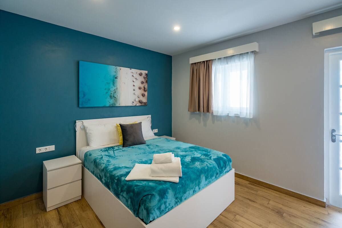 A modern bedroom with a teal accent wall, a large bed with a blue and white bedspread, a white bedside table, and a window with sheer curtains.