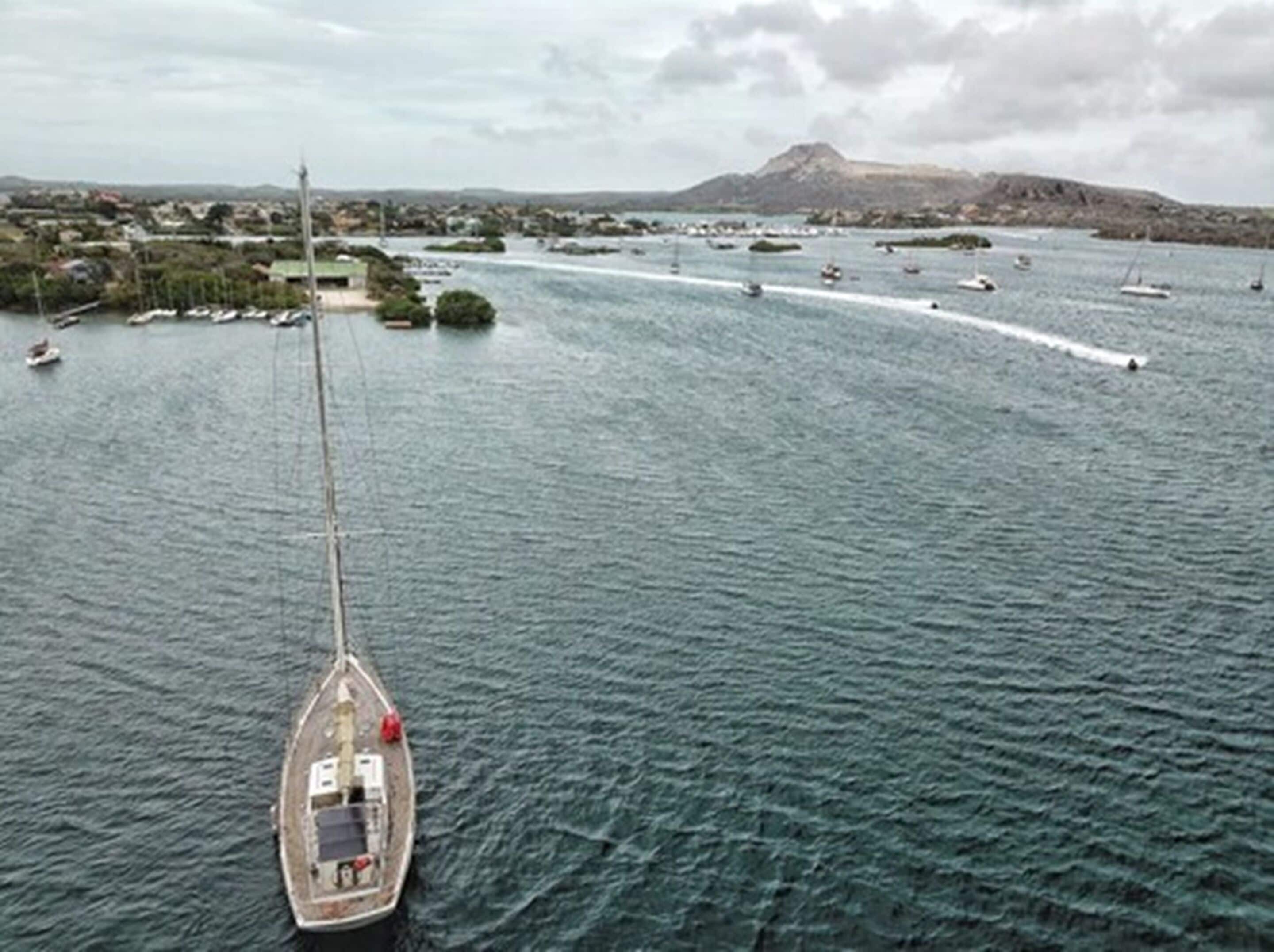 Un velero en un mar tranquilo con montañas al fondo y un pueblo pequeño al otro lado del agua.