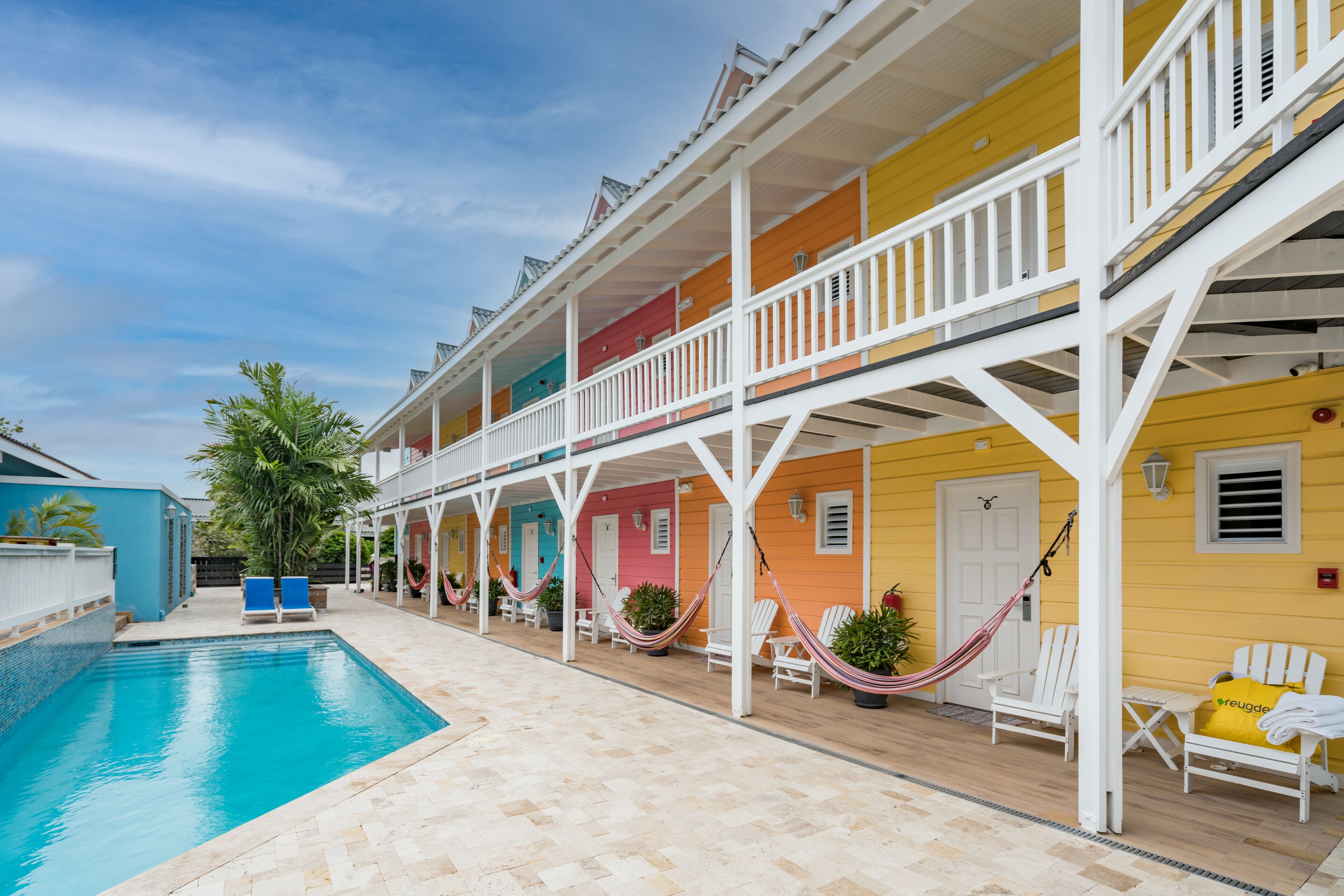 Uma área à beira da piscina com piscina, espreguiçadeiras brancas e edifícios coloridos.