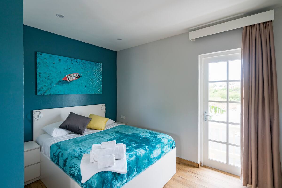 A modern bedroom with a large bed, a teal wall, a window with curtains, and a painting of a boat.