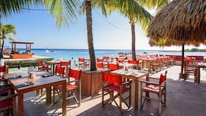 An outdoor dining area with wooden tables and red chairs, overlooking a beach with palm trees and a thatched-roof structure. - Bed & Bike Curacao - Jan Thiel (Jan Thiel)
